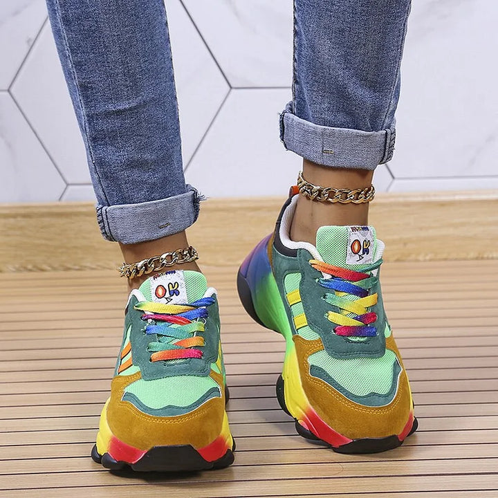Damesmodel draagt de orthopedische regenboogschoenen, zittend, waarbij het comfort en de stijl benadrukt worden.