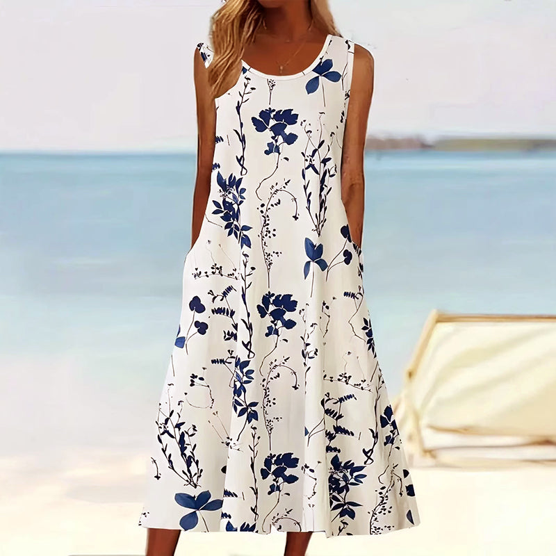 Model draagt een witte mouwloze midi jurk met blauw bloemenpatroon op het strand, zijzakken zichtbaar.