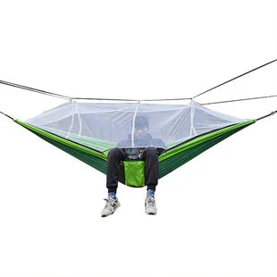 Outdoor hangmat met muskietennet, gemaakt van groene parachutestof, hangend tussen twee bomen, ideaal voor kamperen.